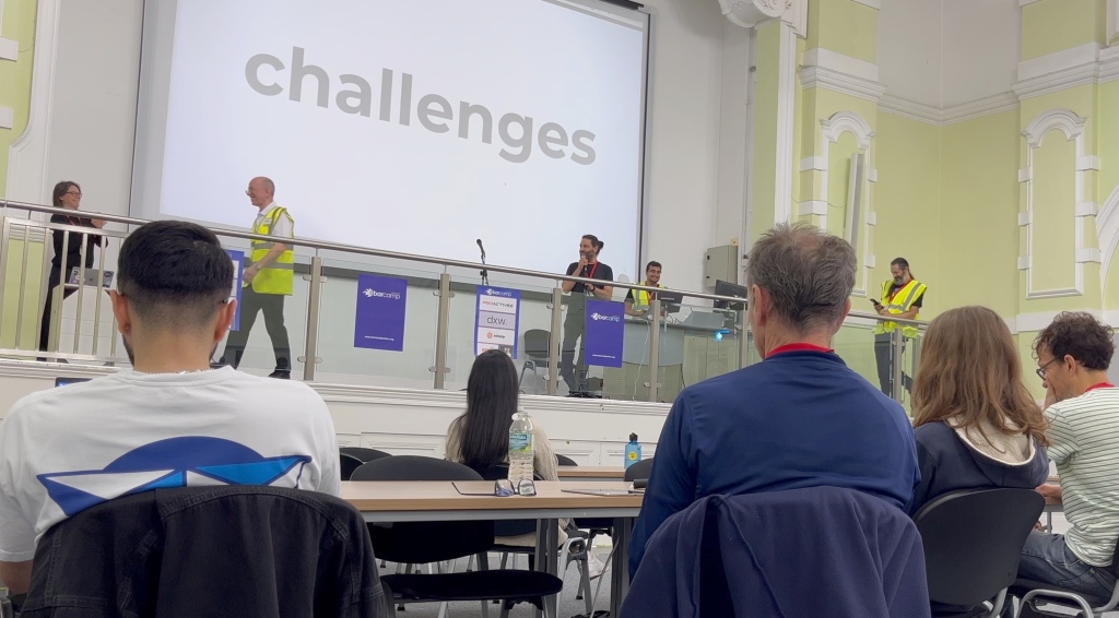 Louie Christie on a stage holding a microphone. A screen behind him says 'challenges'. Several people are on stage laughing at something. Audience of geeks in the foreground.
